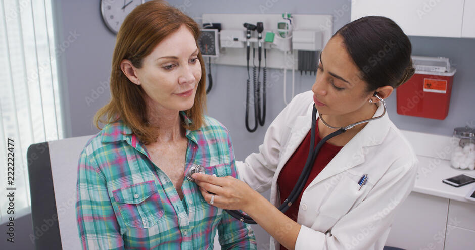 Lovely young latina doctor using stethoscope to examine senior patients vitals. Charming hispanic doctor using medical instrument to check heart and lungs of female caucasian senior patient фотография Stock | Adobe Stock